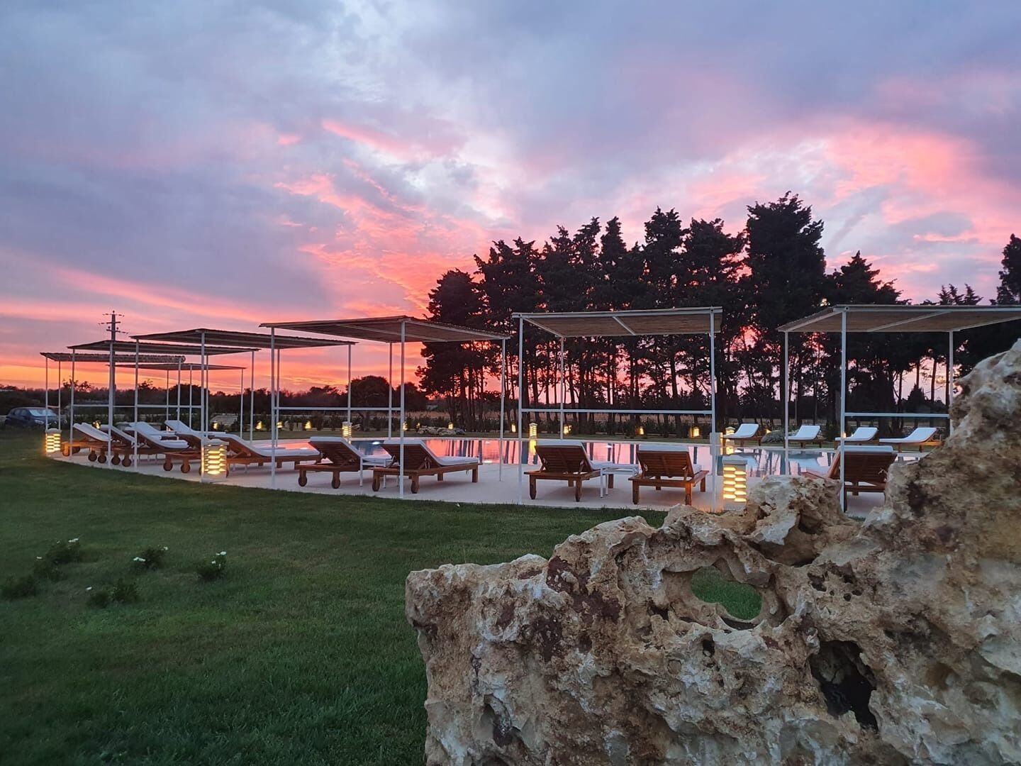 A serene outdoor seating area with lounge chairs and umbrellas, set against a backdrop of trees and a sunset sky.