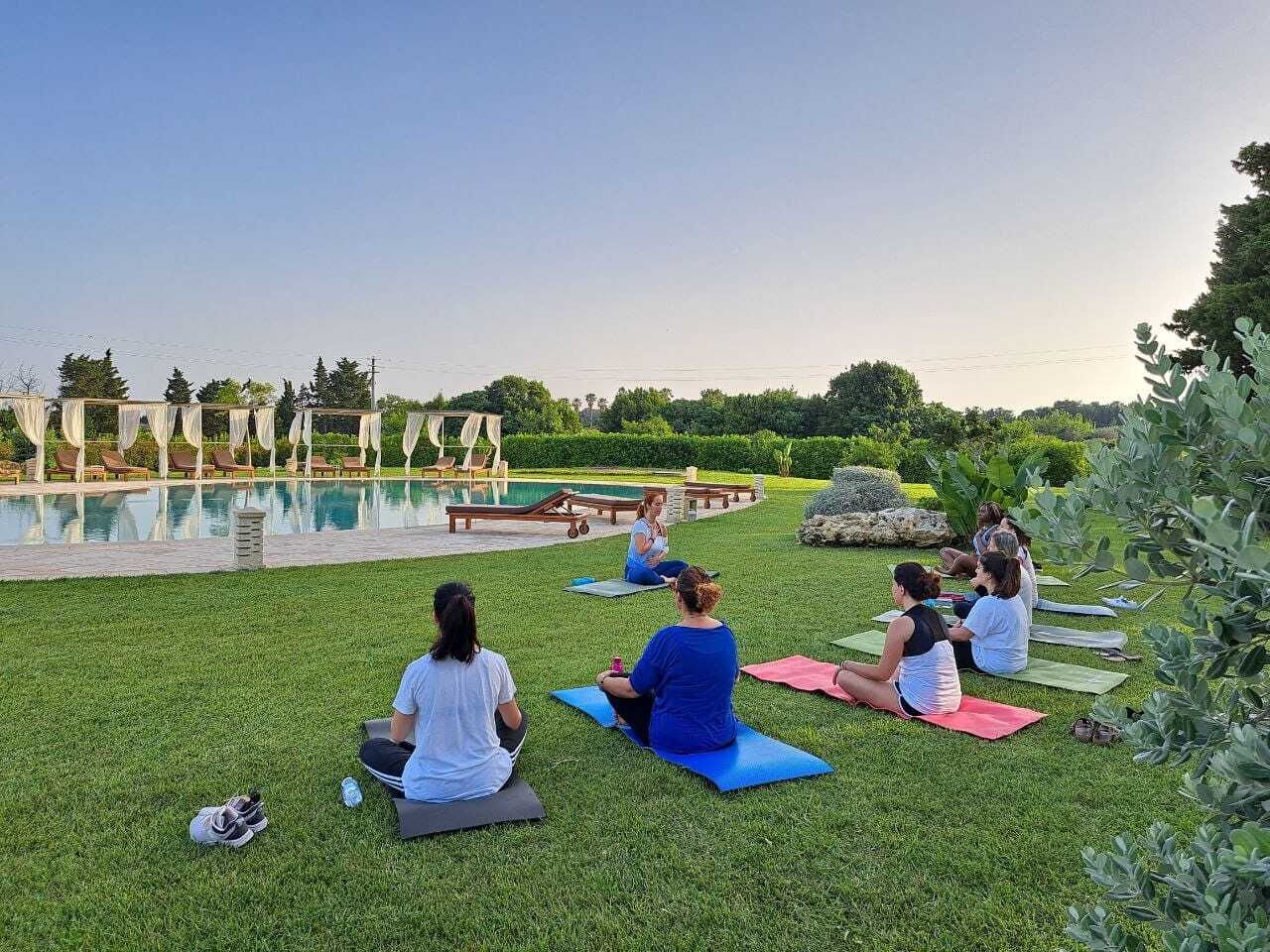A group of people sitting on yoga mats outdoors with a pool and lounge chairs in the background.