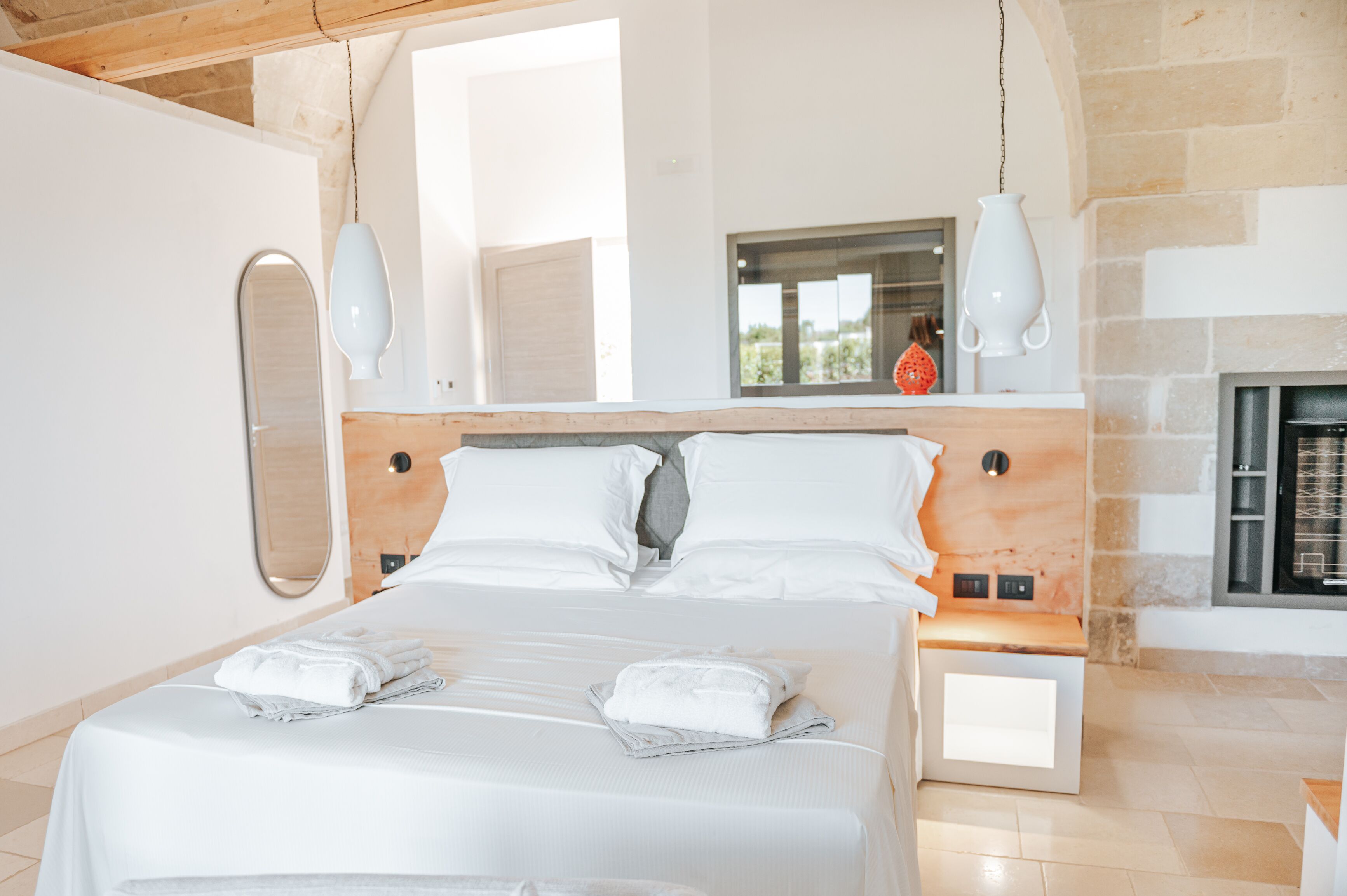 A modern bedroom with a large bed, white linens, a stone fireplace, and hanging pendant lights.