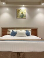 Una cama bien hecha con sábanas blancas y almohadas azules, un cabecero de madera y un cuadro de flores en la pared.
