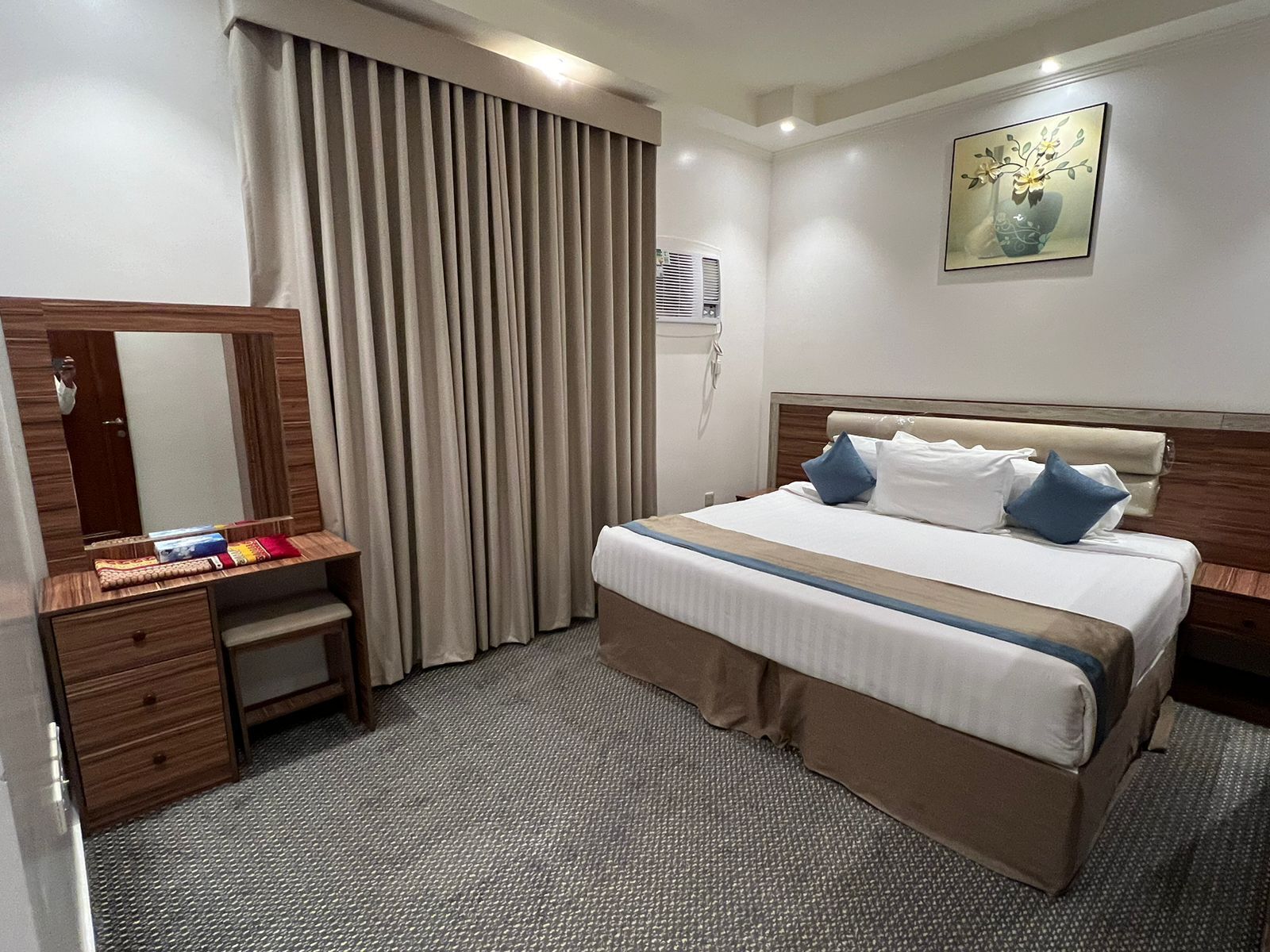 A hotel room with a bed, a desk with drawers, a mirror, a window with curtains, an air conditioning unit, and a painting on the wall.