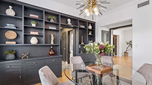 A modern dining area with a glass table, grey upholstered chairs, and a dark wood cabinet with shelves.