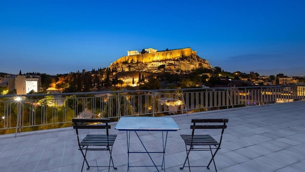 Terraço com mesa e cadeiras dando vista para um edifício histórico no topo de uma colina ao entardecer.