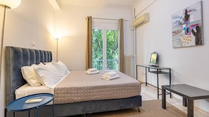 Um quarto com cama, mesas de cabeceira, abajur, quadro na parede e janela com cortinas.