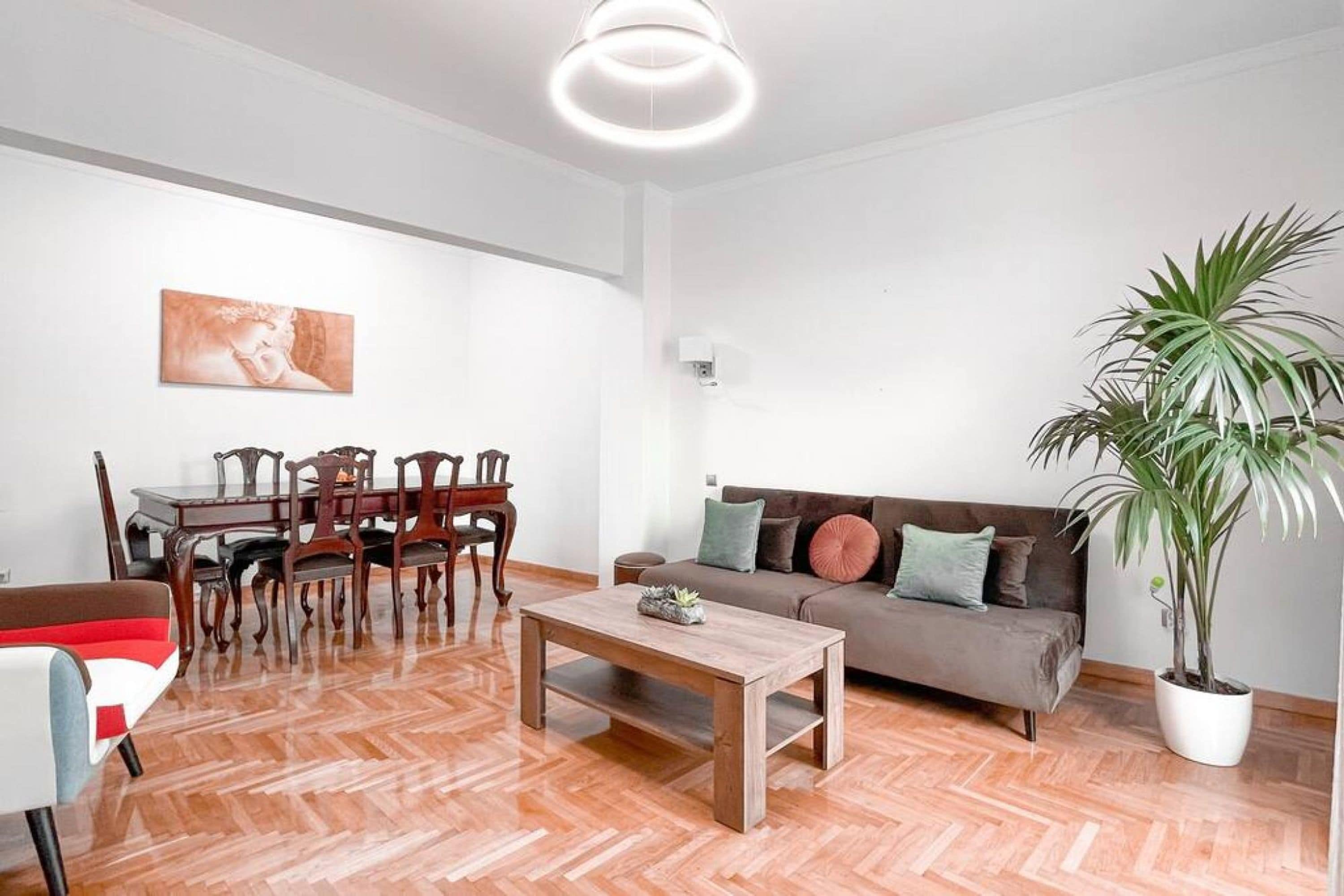 A modern living room with a wooden herringbone floor, a sofa, a coffee table, a dining table with chairs, and a potted plant.