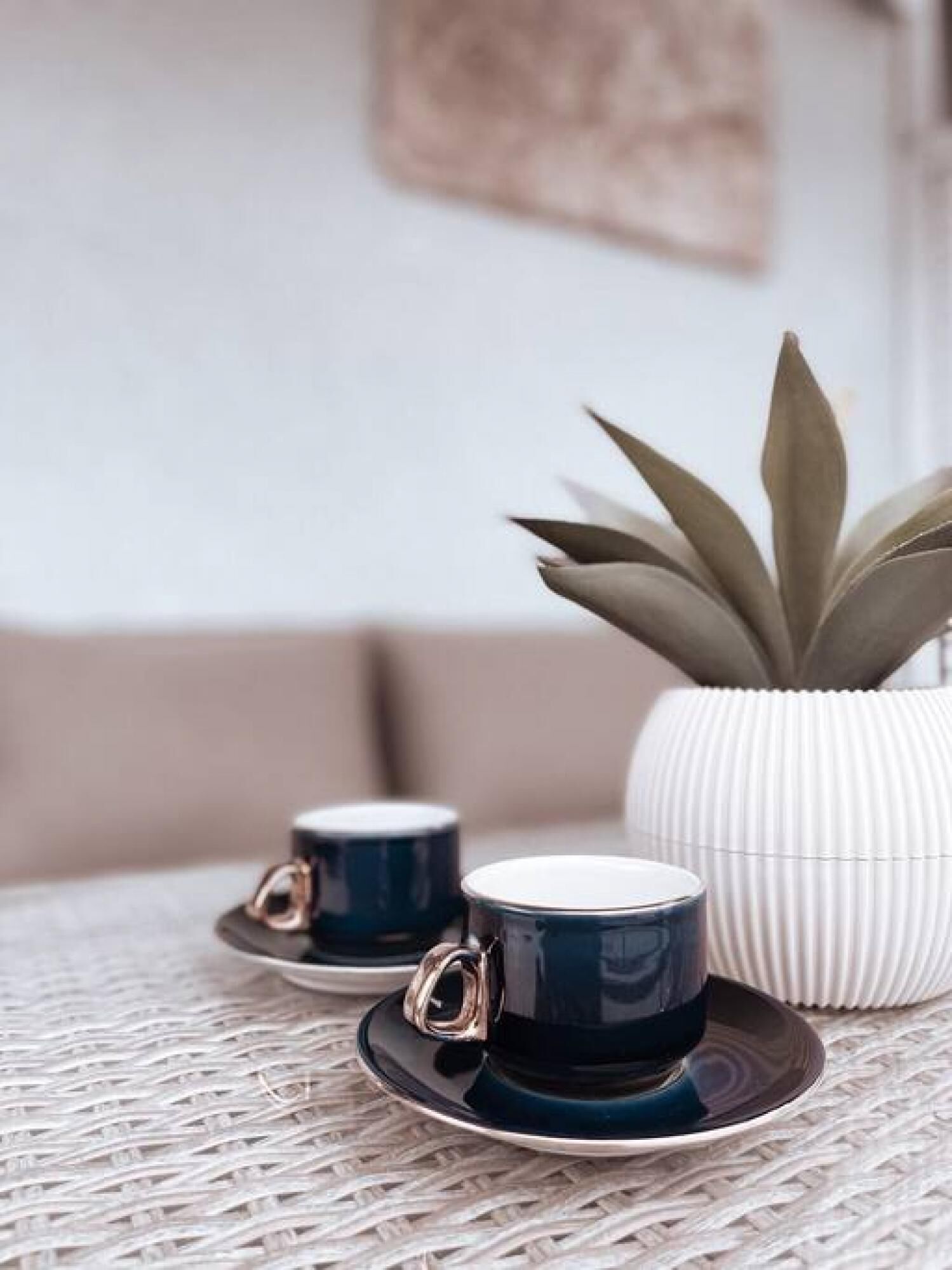 A wicker table with two dark blue cups and saucers, a white ribbed pot, and a green plant in the background.