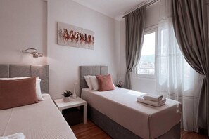 A modern hotel room with two beds, a small bedside table with a plant, a painting on the wall, and a view of the city through a window with curtains. - Penthouse - Amazing View in the Center of Athens (Athens)