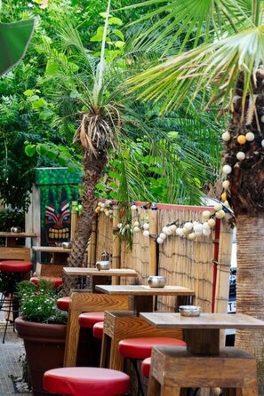 An outdoor dining area with wooden tables and red cushioned benches, surrounded by lush greenery and a bamboo fence. - Penthouse - Amazing View in the Center of Athens (Athens)