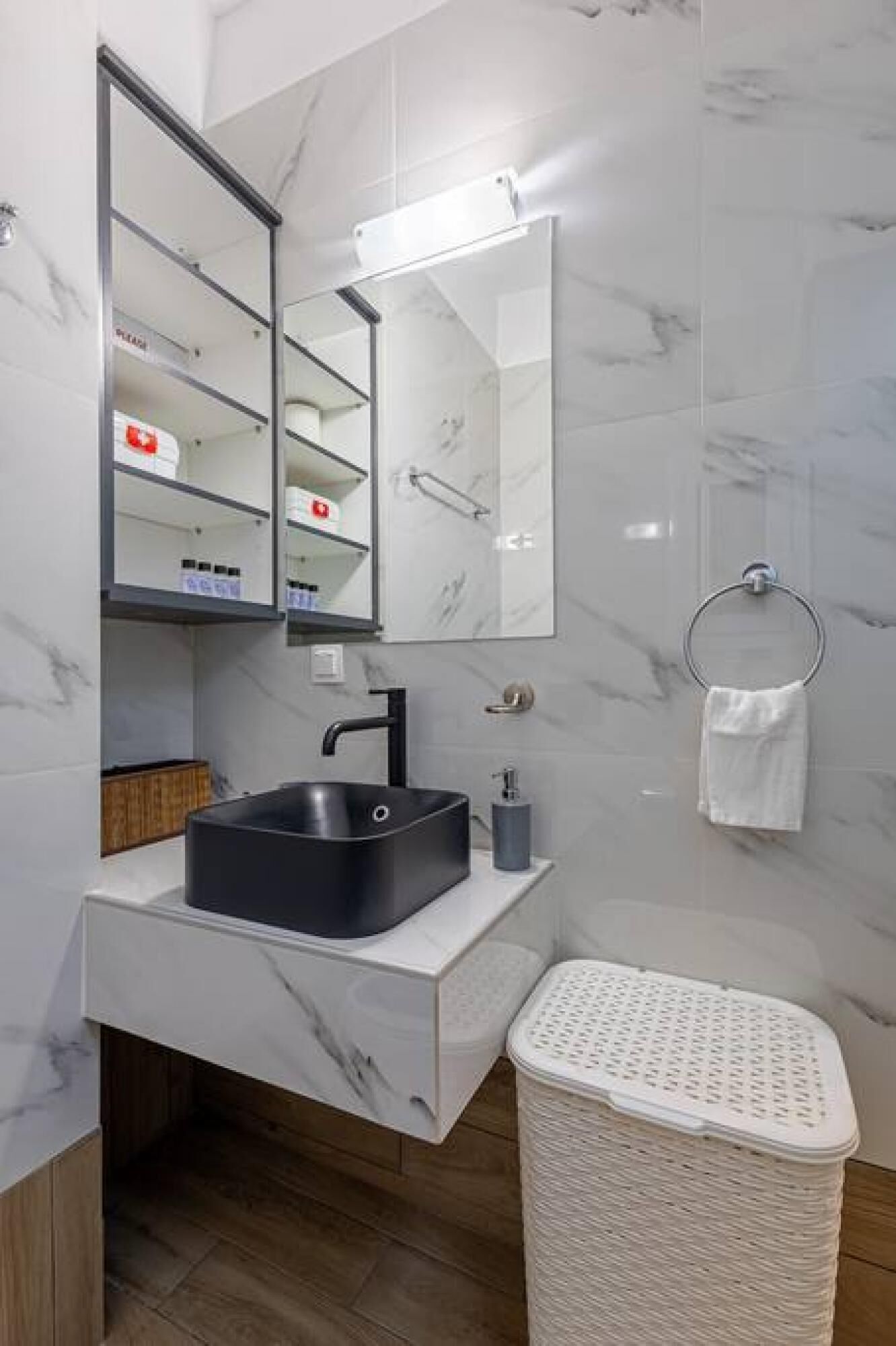 Uma casa de banho moderna com lavatório preto, paredes de mármore branco, um espelho grande e uma cesto de roupa branca.