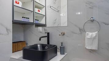 Uma casa de banho moderna com lavatório preto, paredes de mármore branco, um espelho grande e uma cesto de roupa branca.