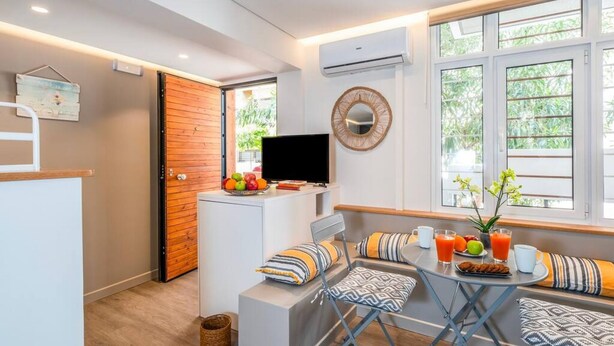 A compact living space with a kitchenette, a dining area with a table and chairs, a TV, and a large window with a view of greenery. - Cozy Nest at Acropolis Footsteps (Athens)