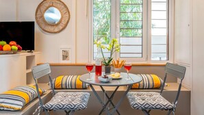 A compact living space with a round table, two chairs, a television, a decorative mirror, and a window with a view of greenery. - Cozy Nest at Acropolis Footsteps (Athens)