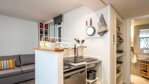 A compact kitchen with a built-in oven, a sink, and a stove. There is a gray sofa in the living area and a small dining table. - Cozy Nest at Acropolis Footsteps (Athens)