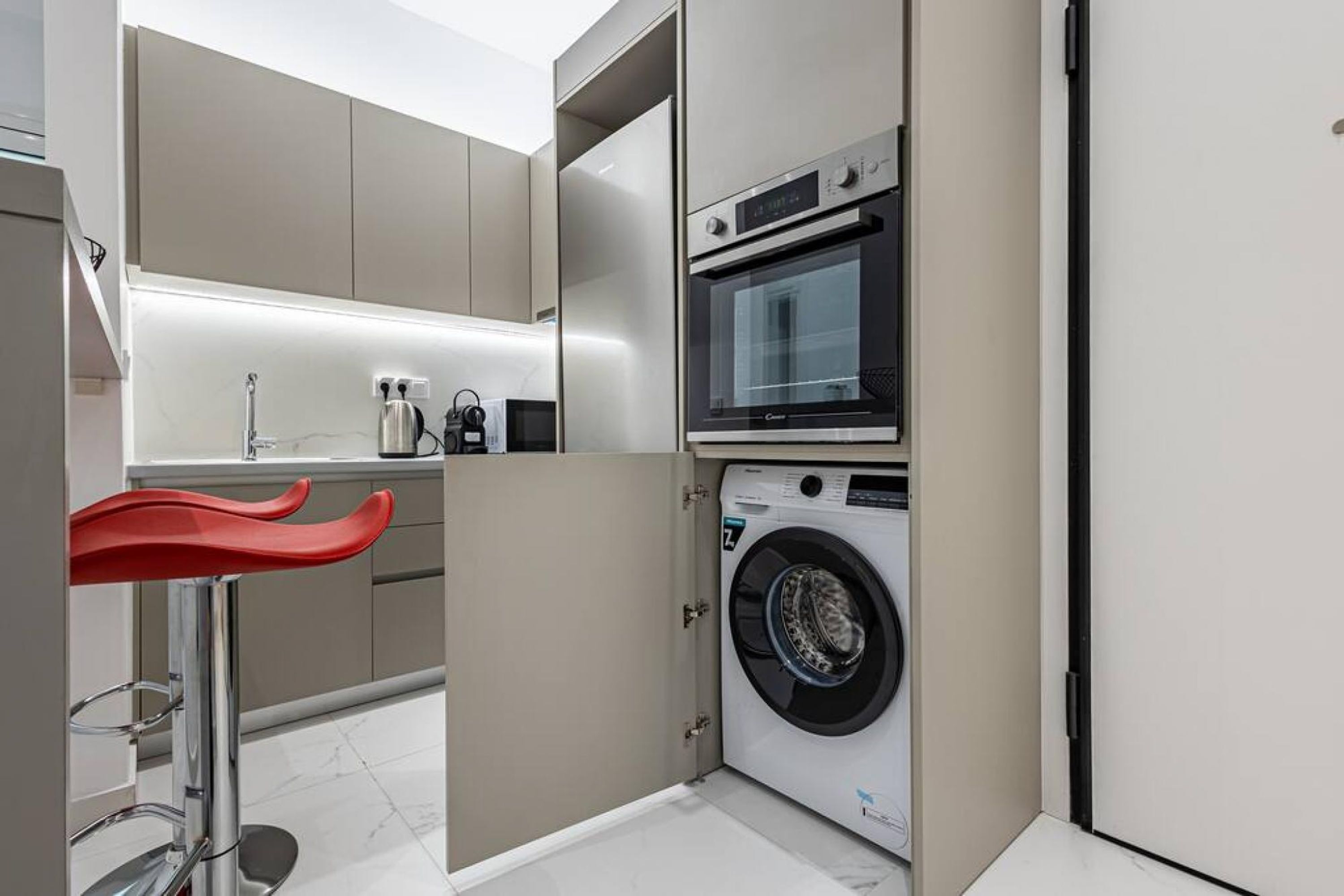 A compact kitchen with a built-in washing machine and oven, a sink, and a red bar stool.