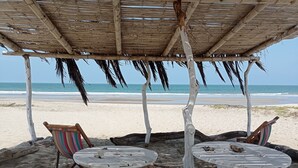 Un espace aménagé avec des sièges, situé au bord de la plage, comprenant deux tables blanches et des chaises à rayures, le tout abrité par un toit de chaume.