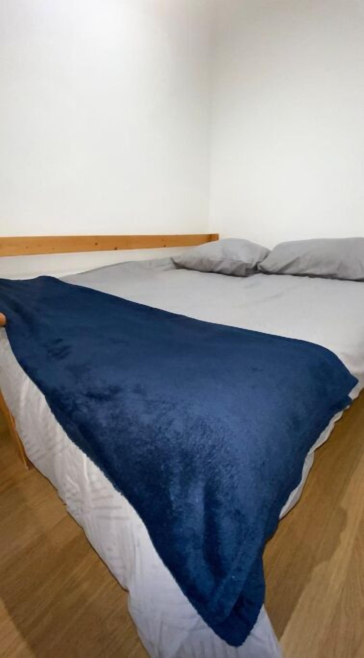 Een bed met een blauwe deken en witte lakens in een kamer met houten vloer en witte muren.