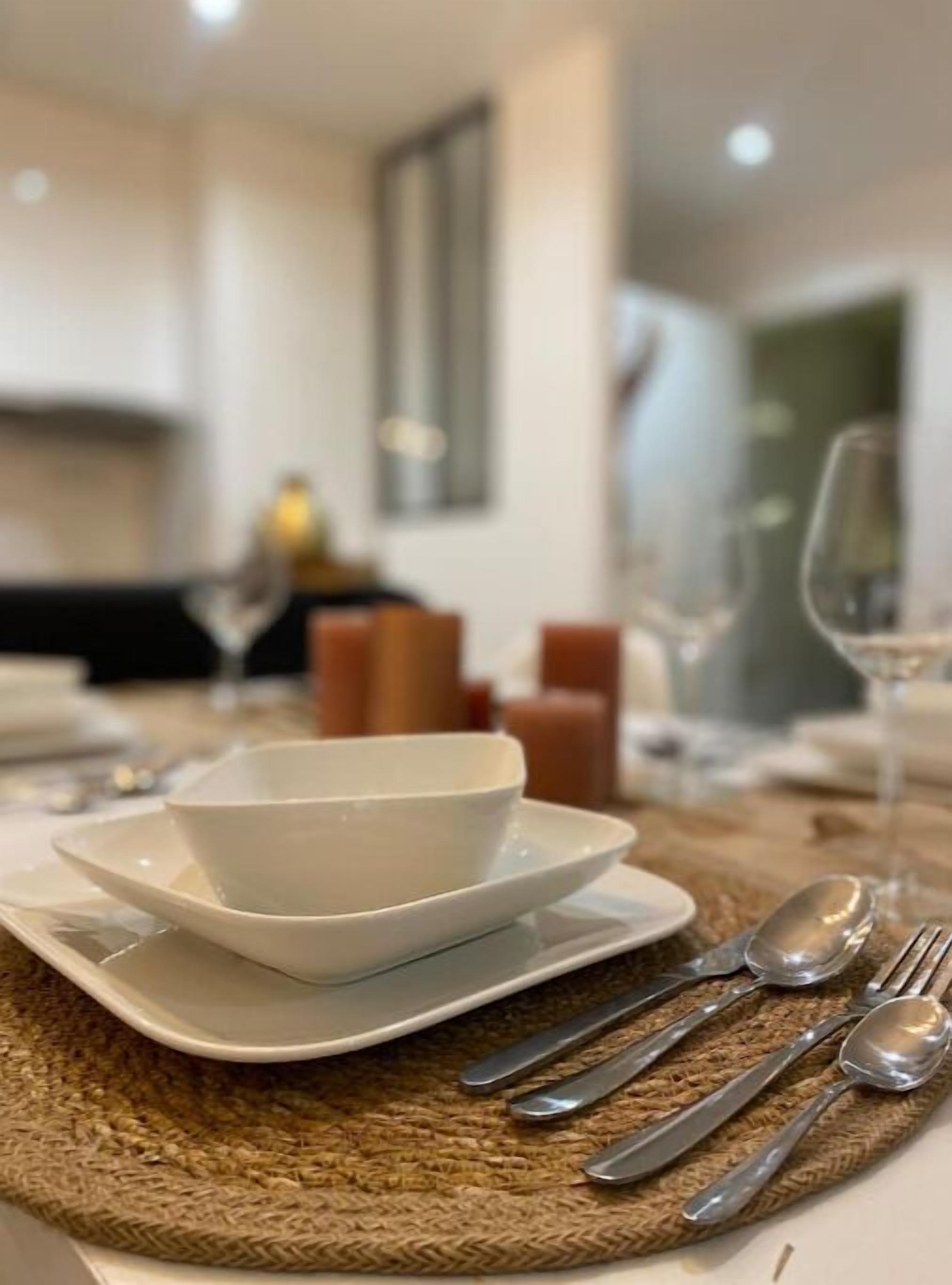 Une table à manger dressée avec de l’assiette blanche, des bols et des couverts en argent, et un set de table en fibres naturelles placé en dessous.