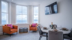 A modern dining area with a round table, two orange armchairs, and a large window offering a view of the sea. - Caldey View - Luxury 2 Bedroom - Panorama - Tenby (Tenby)
