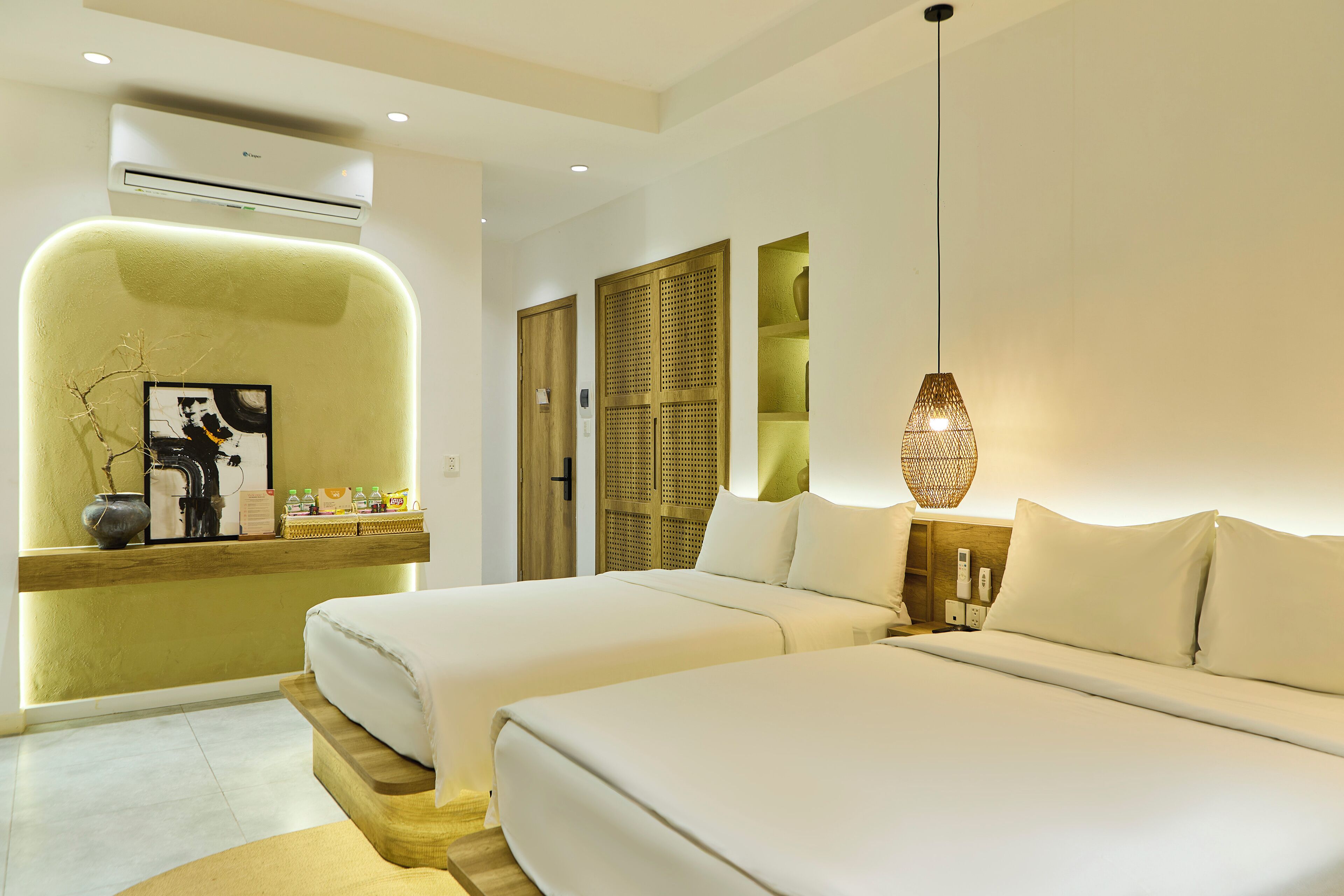 A hotel room with two beds, a wooden headboard, a wall-mounted air conditioner, a shelf with toiletries, and a pendant light.