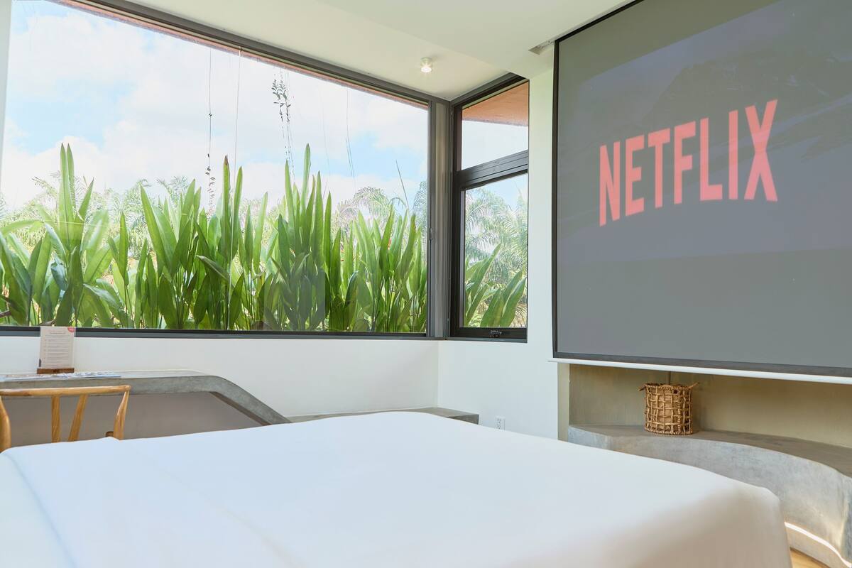 A modern hotel room with a large window, a bed, a small table, and a wall-mounted screen displaying "NETFLIX".
