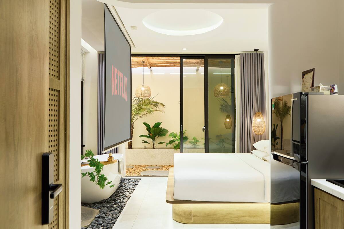 A modern hotel room with a large bed, a flat-screen TV, a minibar, and a sliding glass door leading to a balcony with plants.