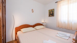 A double bed with a wooden headboard, a bedside table with a lamp, a painting on the wall, and a window with sheer curtains. - Nice apartment on the first floor with a covered balcony (Baska)