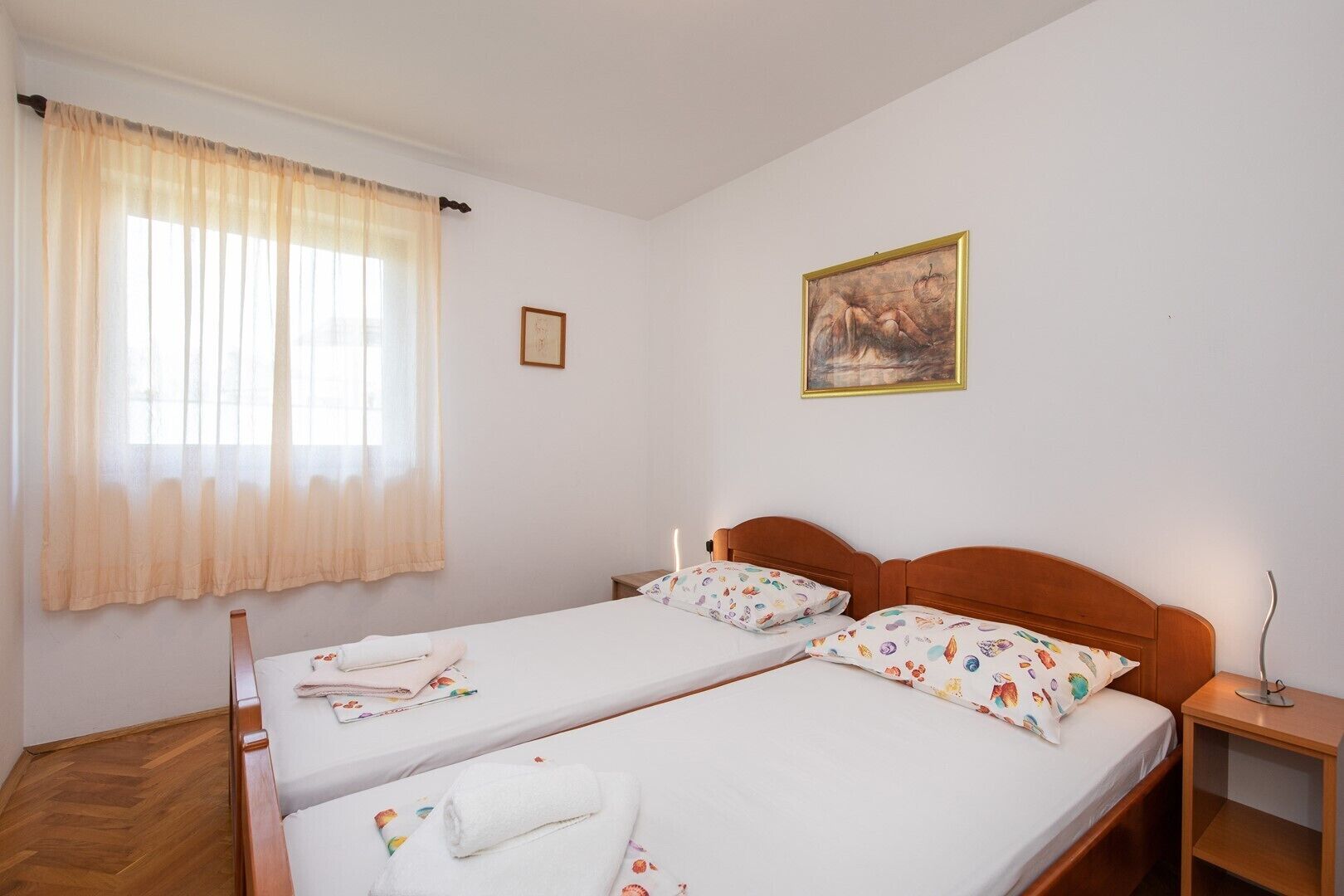 Un lit double avec une literie à motifs floraux, une table de chevet, une commode, une fenêtre avec des rideaux légers et un tableau encadré au mur.