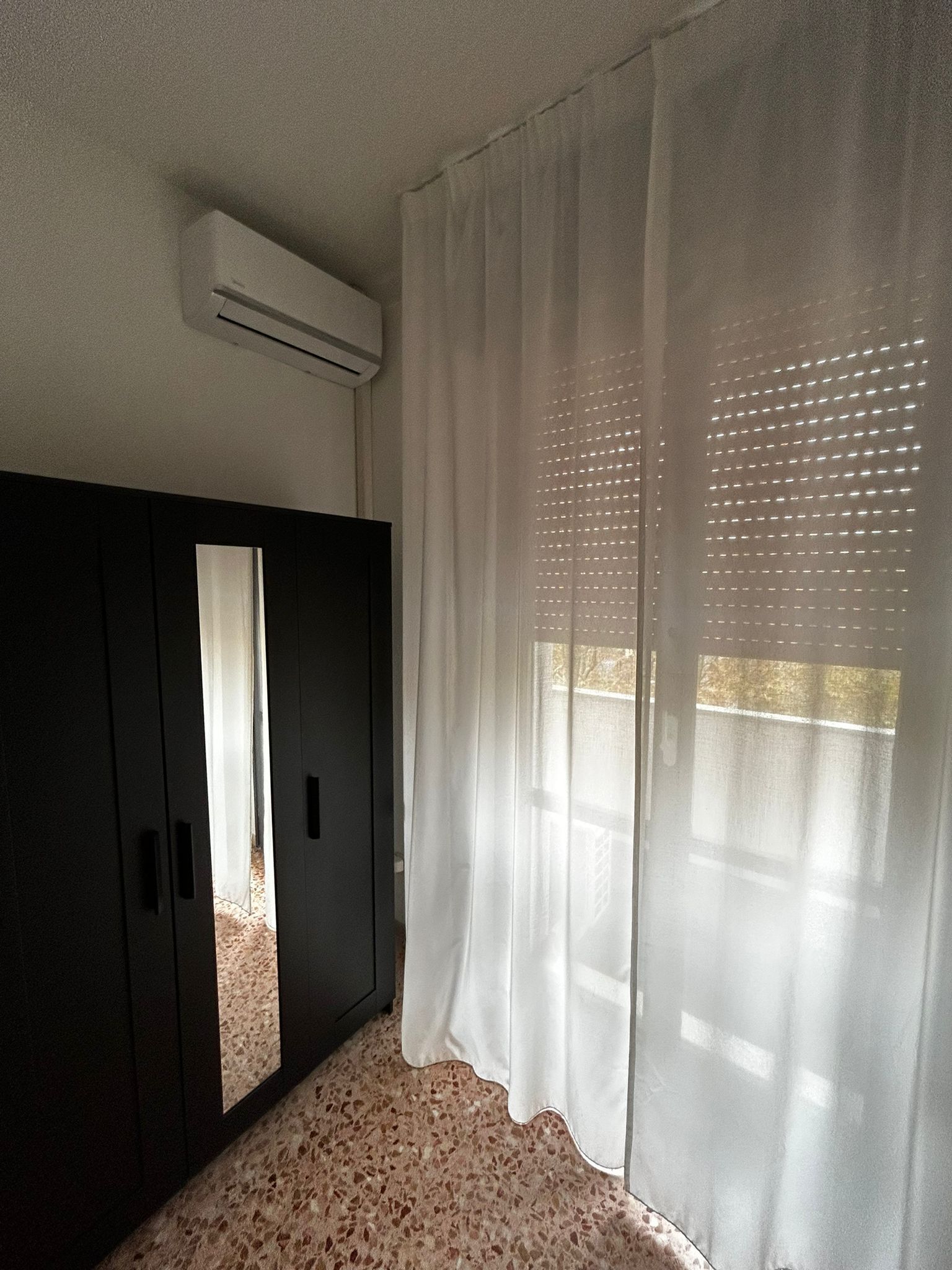 Una habitación con un armario oscuro, un ventanal con cortinas translúcidas y aire acondicionado en el techo.