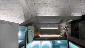 A modern indoor pool with clear glass walls, a wooden deck, and a stone wall in the background. - Palazzo Giusti Suites and Spa (Urbino)