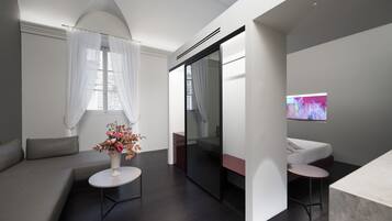Ein modernes Hotelzimmer mit einer Couch, einem kleinen runden Tisch mit Blumenvase, einem Bett mit Fernseher und einer Schiebetür.