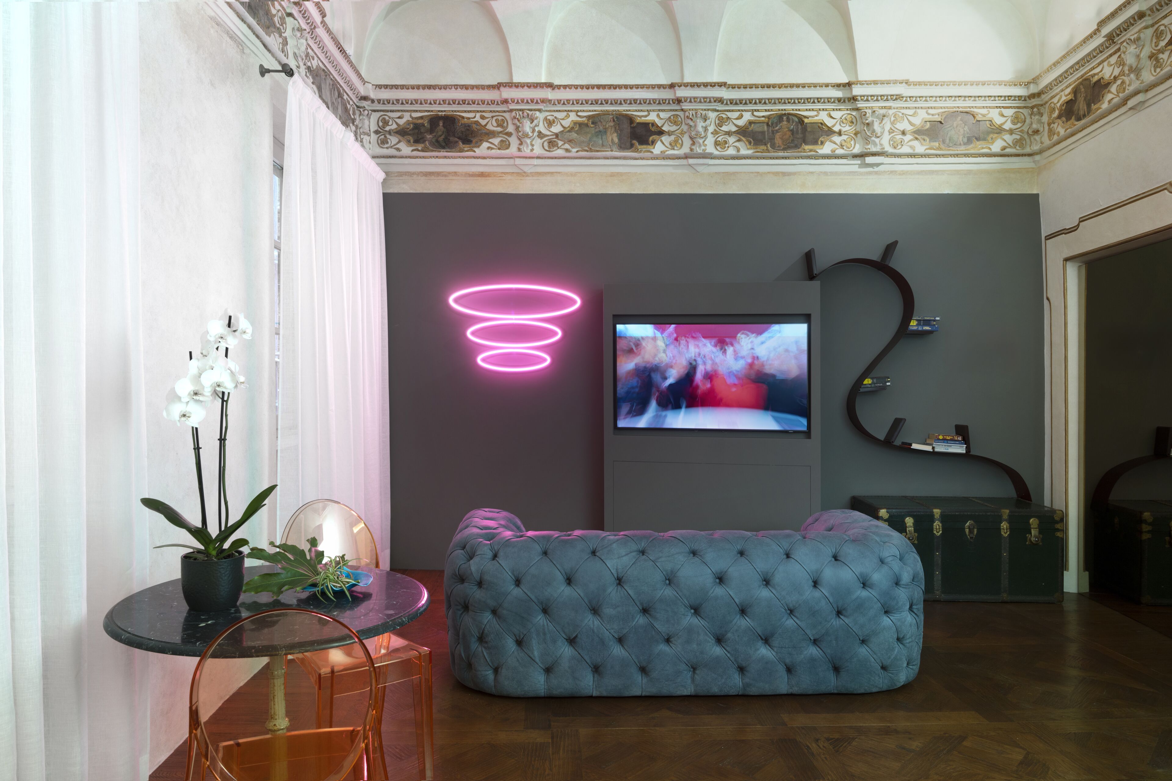 Una sala de estar moderna con un sofá azul capitonado, una mesa redonda de vidrio con sillas, una planta en maceta y una luz de neón.