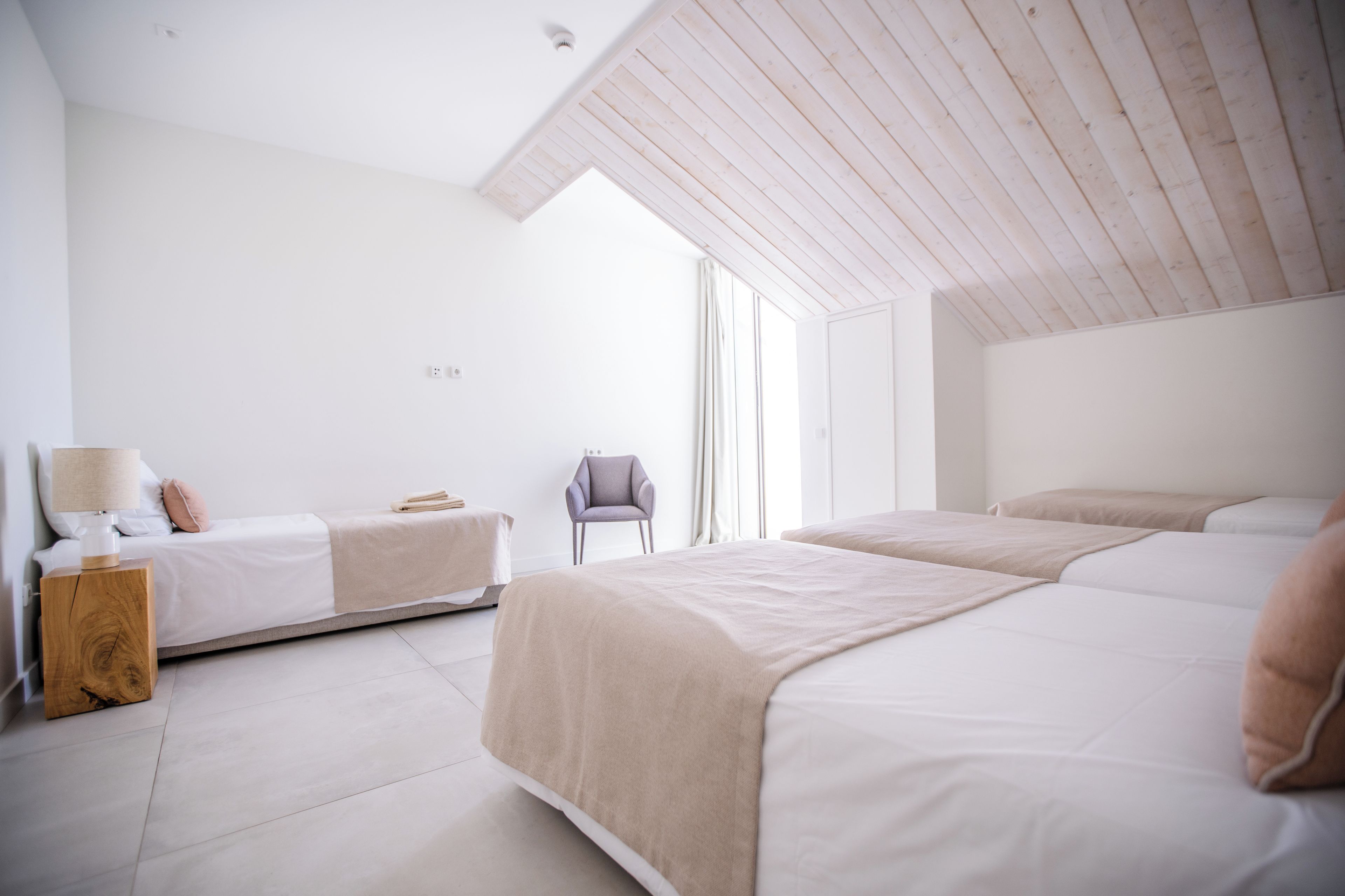Habitación de hotel moderna con techo inclinado, dos camas, una mesita de noche con lámpara y una silla.
