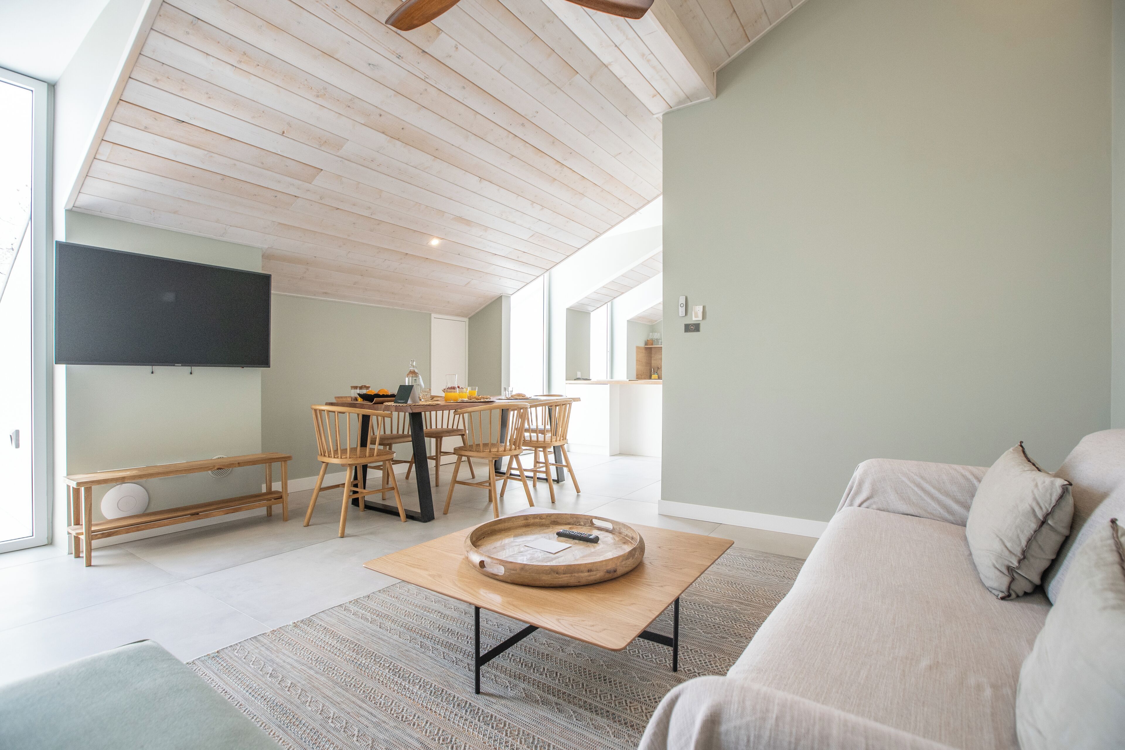 A spacious living room with a high ceiling, a dining area with wooden furniture, and a large flat-screen TV mounted on the wall.