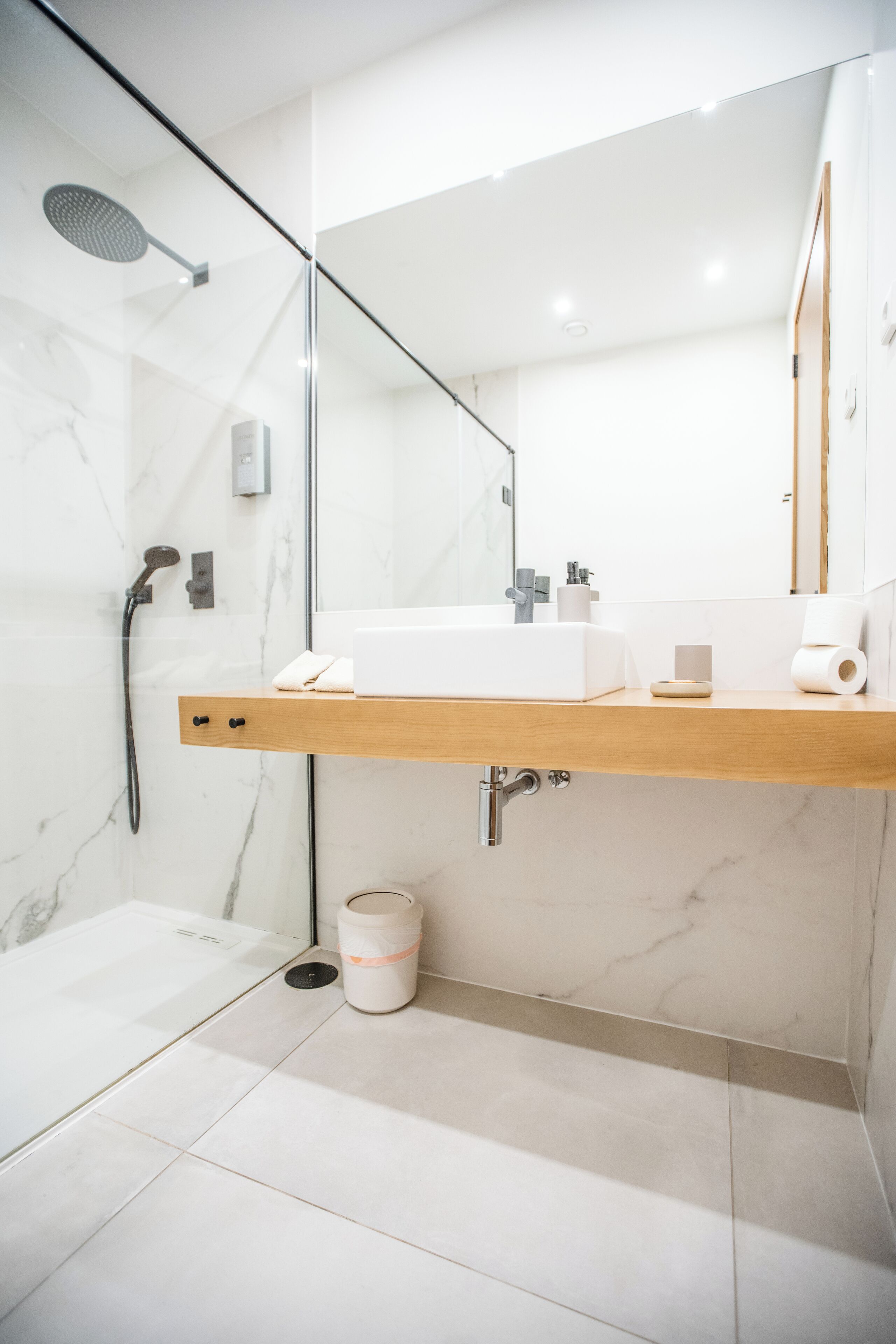 A modern bathroom with a glass shower enclosure, a wooden vanity, a large mirror, and a marble-tiled floor.