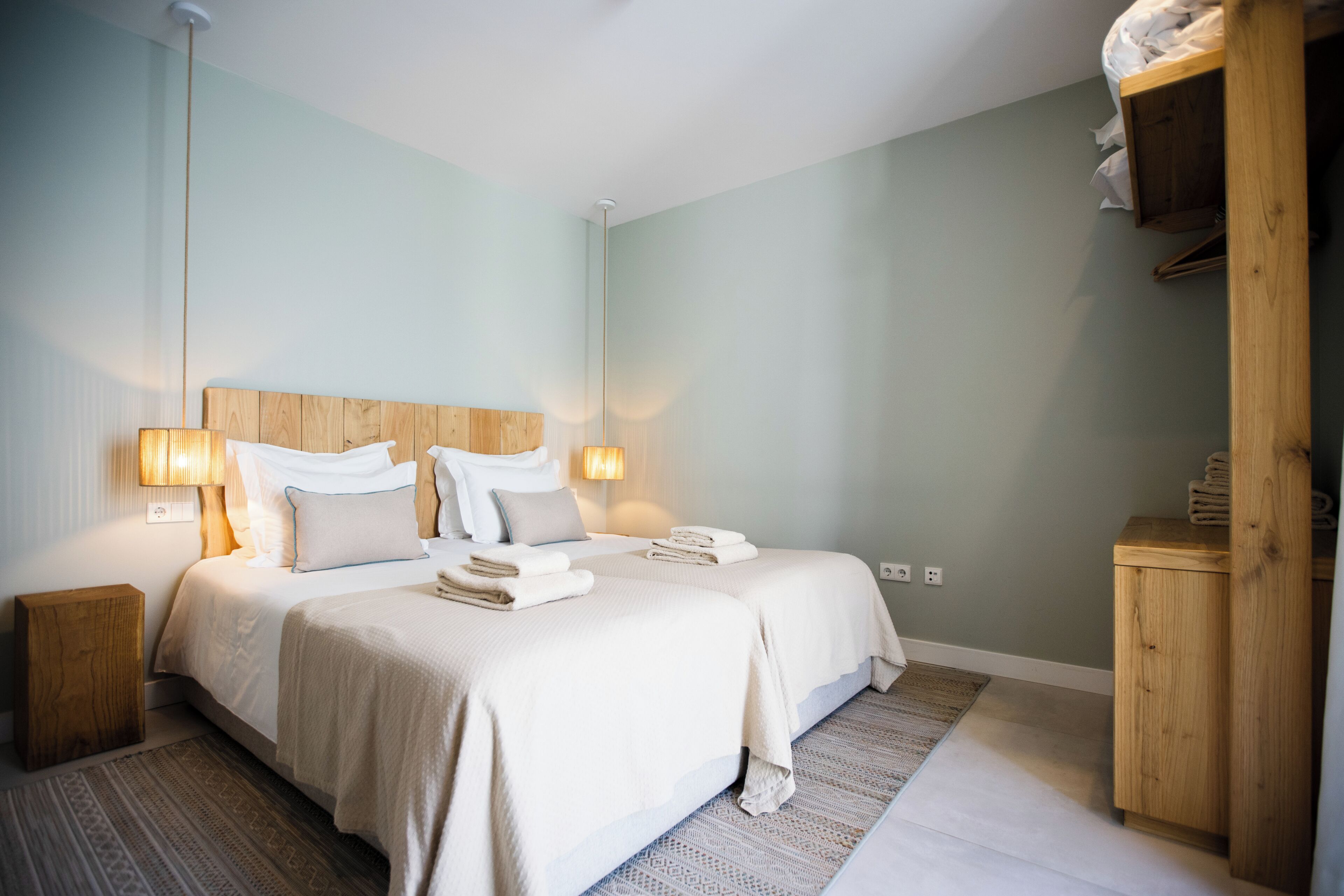 A bedroom with two beds, bedside lamps, a wooden headboard, a nightstand, a patterned rug, and a wooden wardrobe.