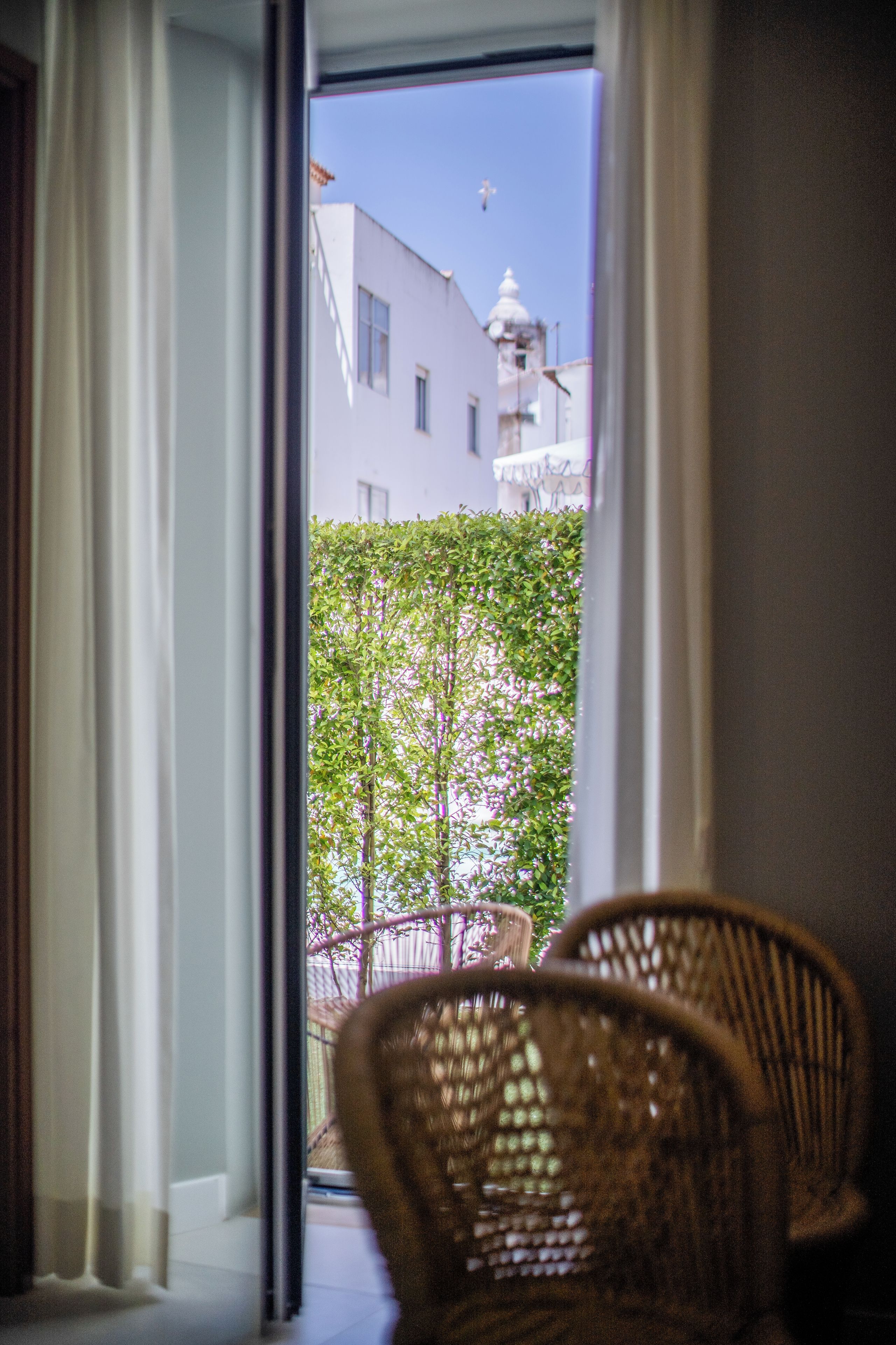 Une vue à travers une fenêtre, avec une chaise en osier au premier plan, un bâtiment blanc surmonté d’un dôme à l’arrière-plan et un ciel dégagé.