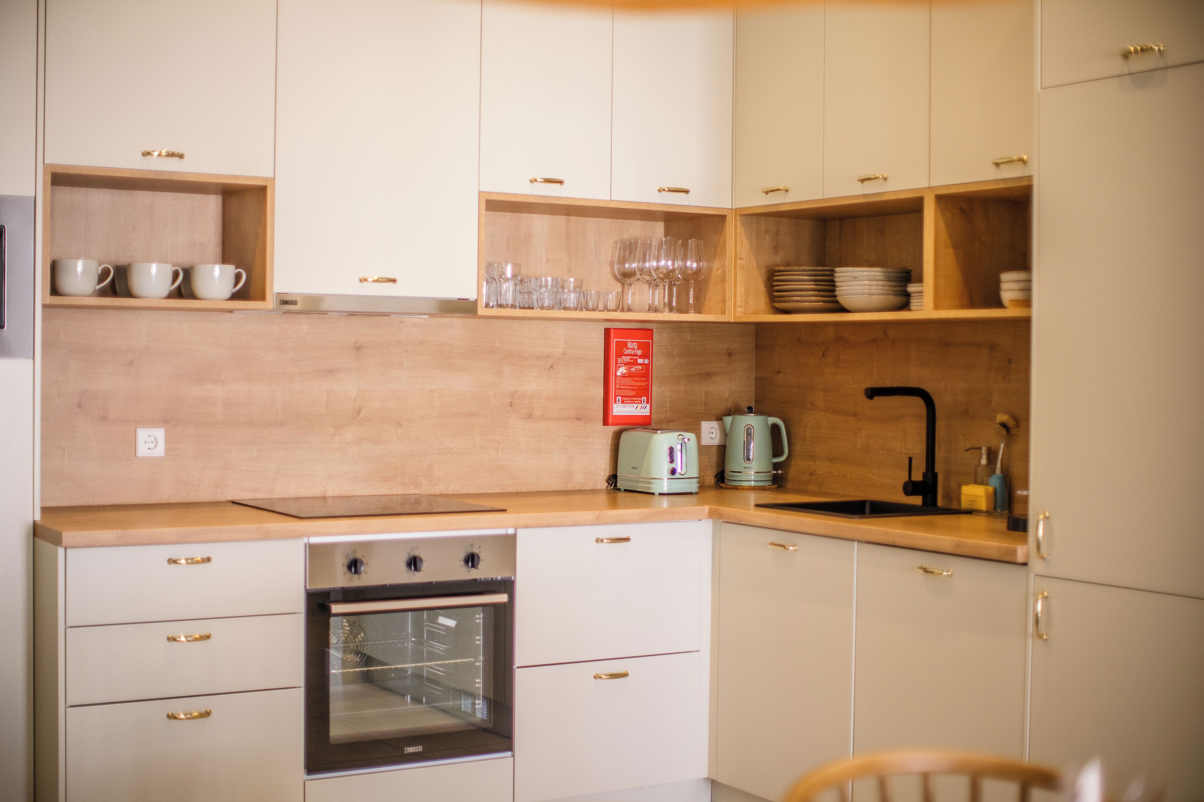 A compact kitchen with white cabinets, a built-in oven, a sink, and wooden shelves holding cups and glasses.