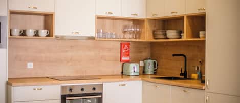 A compact kitchen with white cabinets, a built-in oven, a sink, and wooden shelves holding cups and glasses.
