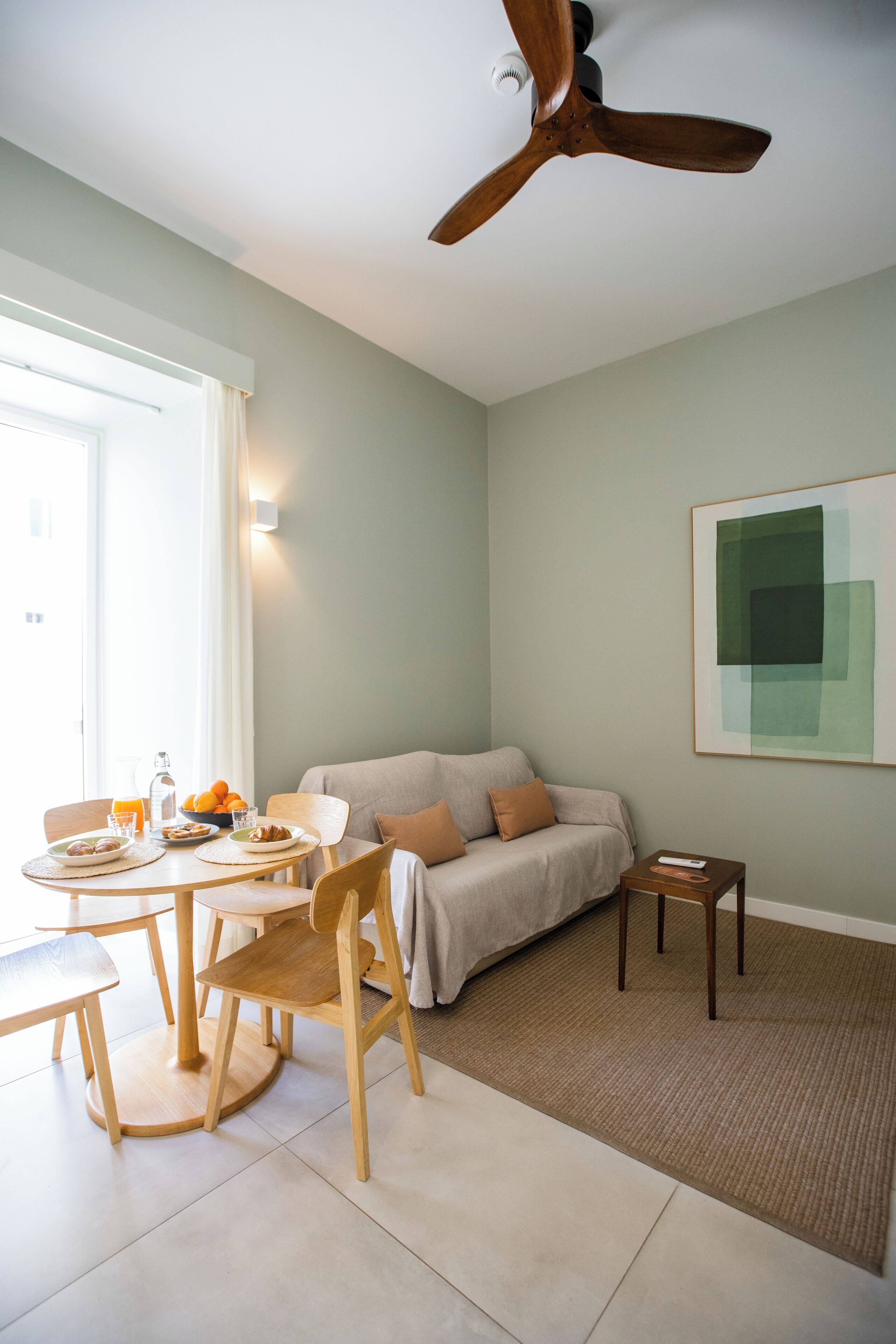 Une pièce avec un ventilateur de plafond, un canapé, une petite table sur laquelle se trouve un saladier rempli de fruits, et un tableau encadré accroché au mur.