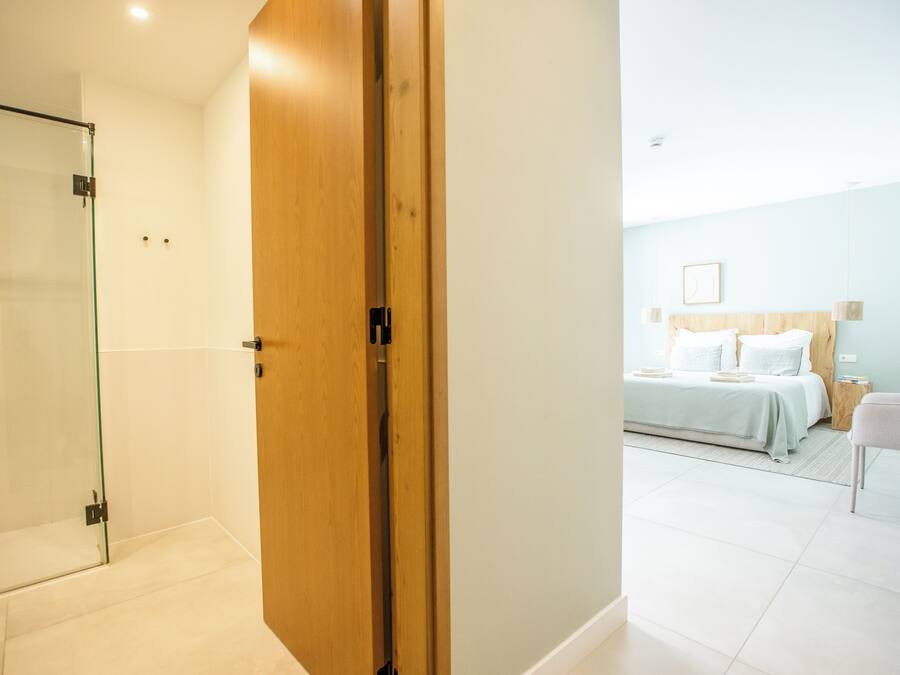 Habitación de hotel con puerta de madera, cabina de ducha de vidrio, cama con ropa blanca y una mesita de noche.