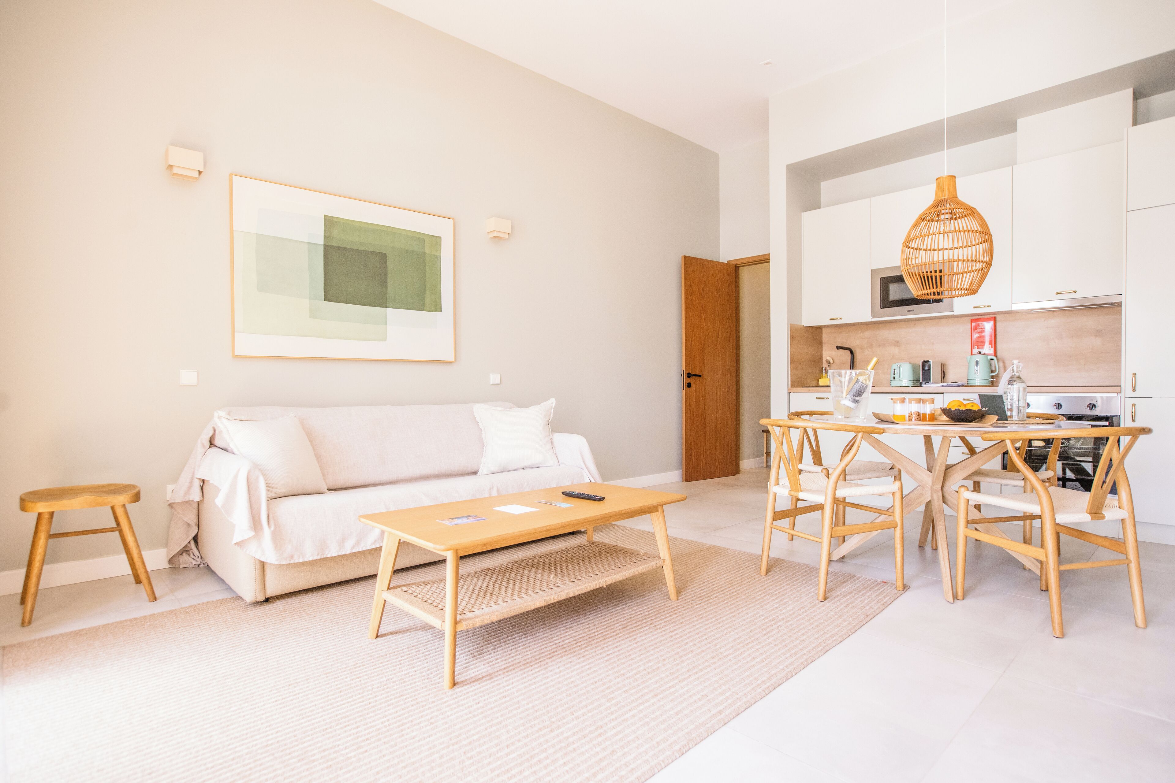 A modern living room with a sofa, coffee table, and dining table with chairs. There is a kitchen area with appliances and a hanging light fixture.