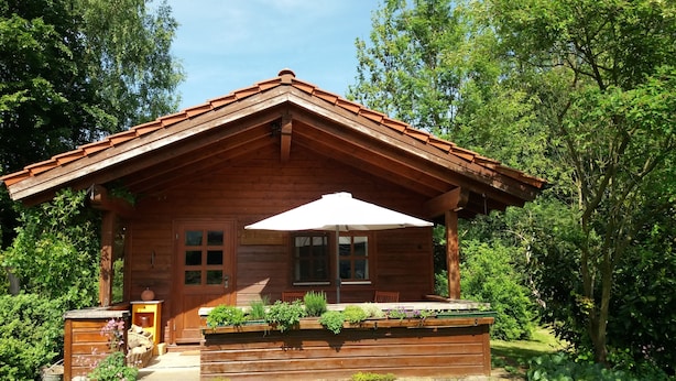 A wooden cabin with a sloped roof, a porch with a bench, and a garden area with plants and flowers.