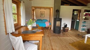 A cozy cabin interior with a wooden dining table, a chair, a bed with blue bedding, and a wood-burning stove.