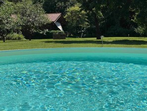 A clear swimming pool surrounded by lush greenery and a building with a red roof. - Romantisches Blockhaus an der Märchenstraße! (Reinhardshagen)
