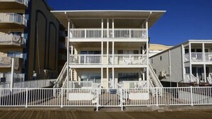 Een meerlaags hotel met balkons, een houten terras op de voorgrond en een helderblauwe lucht.
