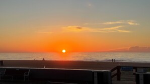 Een zonsondergang boven een strand met een houten promenade, bankjes en een informatiebord.