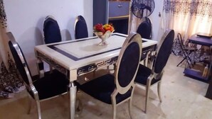 A dining table with a white tablecloth and a bowl of fruit. There are six chairs around the table, and a fan is visible in the background. - Biddex Lodge - by Cenlotts (Benin City)