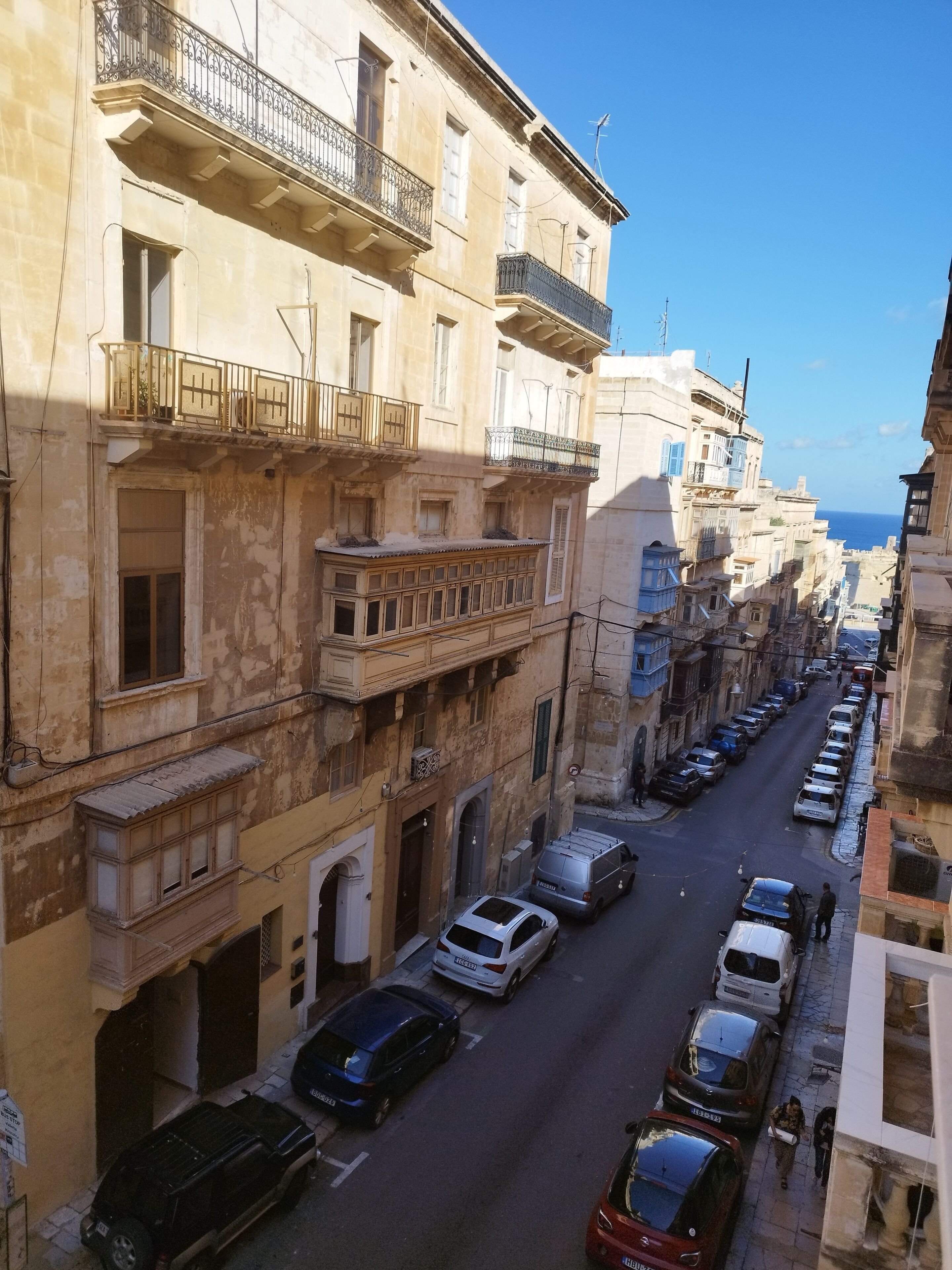 A narrow street lined with multi-story buildings, featuring balconies and a clear view of the sea in the distance.