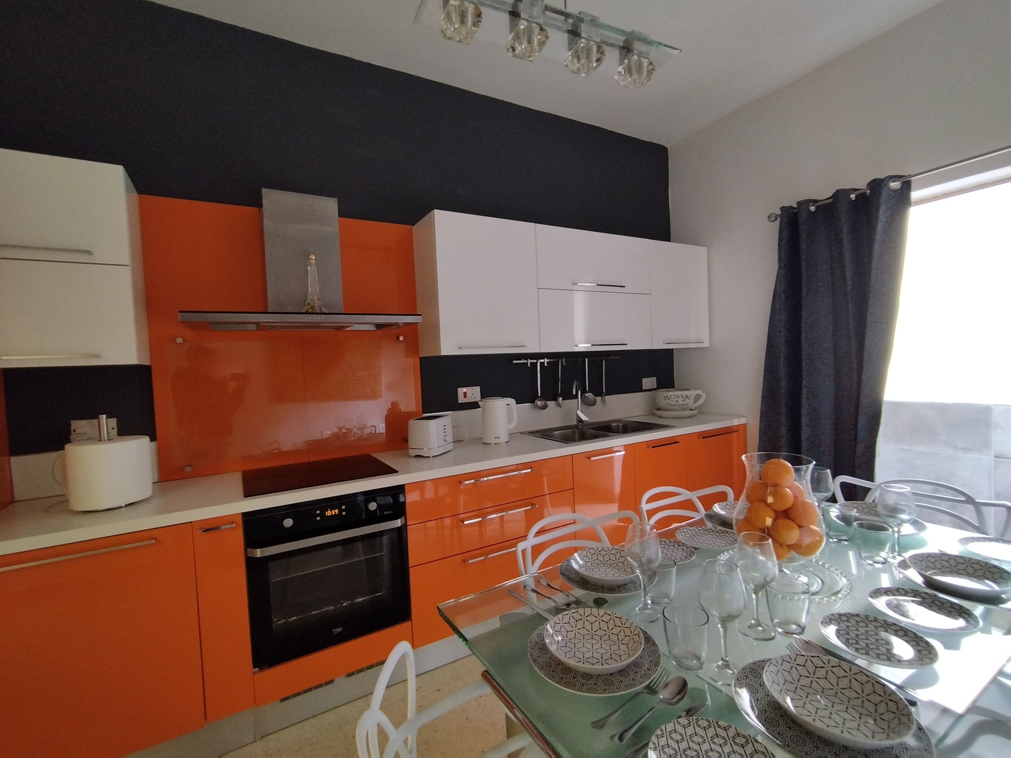 Ett modernt kök med orange skåp, en inbyggd ugn och en matplats med plats för sex personer, med mönstrade tallrikar och glas.
