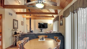 Uma sala de jantar com uma mesa e cadeiras de madeira, um sofá e uma televisão ao fundo.